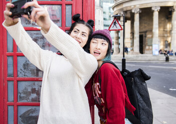 Two young stylish women taking selfie by red phone box, London, UK - ISF03821