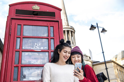 Zwei junge, stilvolle Frauen, die auf ein Smartphone neben einer roten Telefonzelle schauen, London, UK - ISF03823