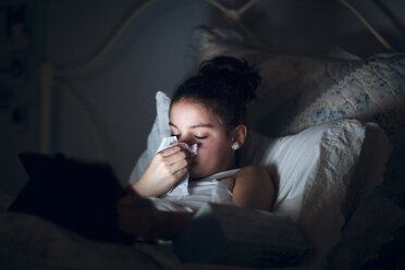 Mädchen im Bett, das sich die Nase putzt, beleuchtet vom Licht eines digitalen Tablets - ISF03837