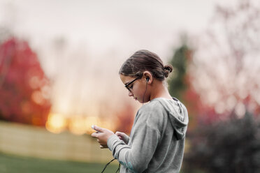 Side view of girl listening to earphone music looking at smartphone in garden - ISF03866