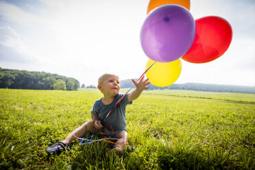 Baby boy sitting in rural field with bunch of colourful balloons - ISF03961