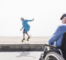 Älterer Mann im Rollstuhl beobachtet seine Frau bei einem Skateboardtrick am Strand, Santa Monica, Kalifornien, USA - ISF04238