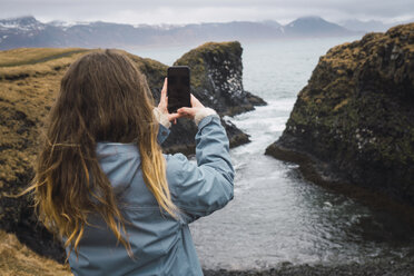 Island, Rückenansicht einer jungen Frau, die an der Küste ein Foto mit ihrem Smartphone macht - KKAF01011