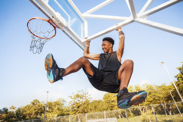 Young man on basketball court, swinging on basketball net frame, low angle view - ISF04574