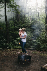 Mutter trägt Tochter und küsst sie auf die Wange am Lagerfeuer im Wald, Huntsville, Kanada - ISF04589