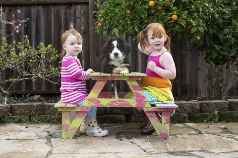 Zwei junge Schwestern sitzen auf einer Gartenbank mit Hund, lizenzfreies Stockfoto