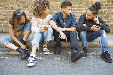 Four friends sitting in street, looking at smartphones - ISF04873