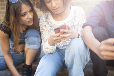 Three friends sitting in street, looking at smartphone - ISF04876