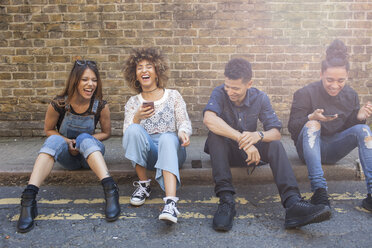 Four friends sitting in street, laughing, young woman holding smartphone - ISF04881