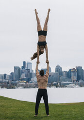Junge Frau balanciert auf den Händen eines Mannes, in voller Länge, übt Yoga vor der Skyline von Seattle - ISF04989