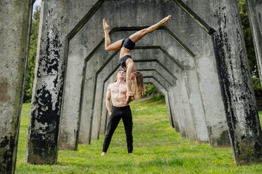 Young woman balancing on top of man hands, practicing yoga below concrete bridge - ISF04990