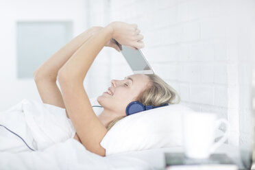 Junge Frau liegt im Bett, trägt Kopfhörer und hält ein digitales Tablet - ISF05036