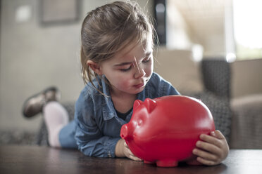 Girl lying on coffee table gazing at red piggy bank - ISF05080