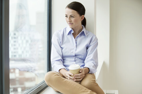 Geschäftsfrau sitzt auf einem Fenstersims, schaut aus dem Fenster und hält eine Tasse Kaffee zum Mitnehmen - ISF05097
