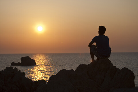 Silhouette eines Mannes auf einem Felsen mit Blick auf den Sonnenuntergang über dem Meer, Olbia, Sardinien, Italien, lizenzfreies Stockfoto