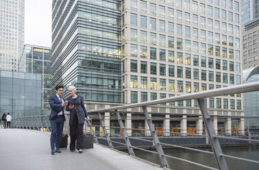 Geschäftsmann und Geschäftsfrau benutzen ein Mobiltelefon und ziehen einen Gepäckwagen, Canary Wharf, London, UK - ISF05602