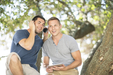 Zwei junge männliche Freunde sitzen unter einem Baum und hören Kopfhörer - ISF05685