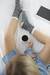 Junge Frau auf dem Bett sitzend, Kaffeetasse in der Hand, digitales Tablet neben ihr, Draufsicht - ISF05714