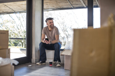 Mann sitzt in einer unmöblierten Wohnung, umgeben von Pappkartons, und benutzt sein Smartphone - ISF05744
