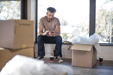 Mann sitzt in einer unmöblierten Wohnung, umgeben von Pappkartons, und benutzt sein Smartphone - ISF05745