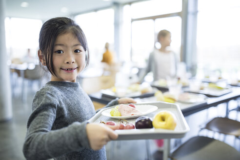 Cheerful schoolgirl holding a tray of food in the cafeteria with a bright smile on her face - WESTF24110