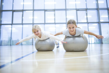 Fröhliche Schulmädchen balancieren auf Gymnastikbällen während eines lustigen Sportunterrichts und zeigen ihr strahlendes Lächeln für die Kamera - WESTF24131