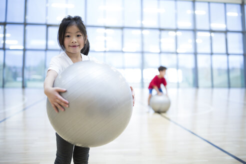 Ein fröhliches Schulmädchen posiert während des Sportunterrichts mit einem Gymnastikball und strahlt dabei Positivität und Begeisterung aus. - WESTF24134