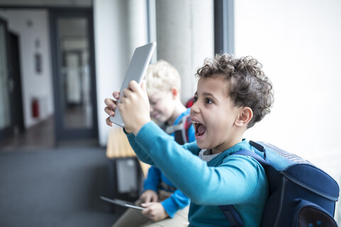 Fröhlicher Schüler zeigt stolz sein Tablet, während er durch den Schulflur läuft, lizenzfreies Stockfoto