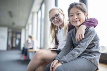 Two happy schoolgirls sitting and smiling in a brightly lit school hallway - WESTF24176