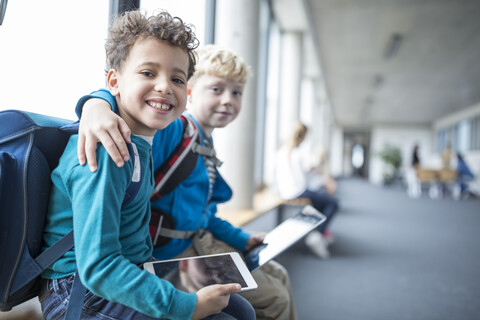 Two happy schoolboys sitting on a bench, embracing each other while holding a digital tablet stock photo