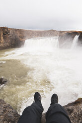 Iceland, legs of a man sitting at Godafoss waterfall - KKAF01033