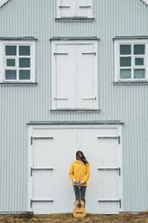 Iceland, woman with guitar standing at house - KKAF01049