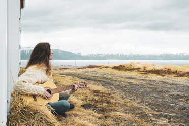 Island, Frau sitzt in ländlicher Landschaft und spielt Gitarre - KKAF01054