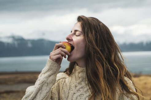 Iceland, woman eating an apple at lakeside - KKAF01057