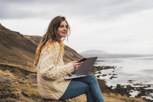 Island, Frau mit Laptop und Mobiltelefon an der Küste - KKAF01069