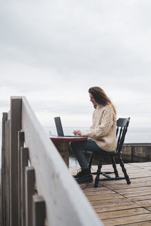 Iceland, woman using laptop at the sea - KKAF01075