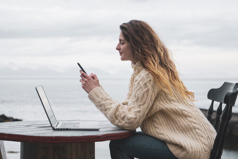 Island, Frau mit Laptop und Mobiltelefon am Meer - KKAF01077