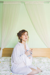 Young woman kneeling on bed holding coffee cup and staring - ISF05910