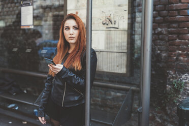 Red haired woman waiting at bus stop holding smartphone - ISF05918