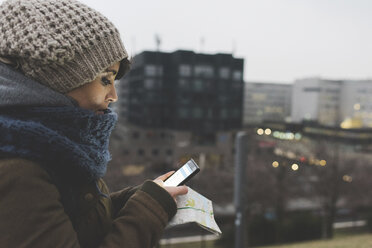 Frau mit Strickmütze schaut in der Abenddämmerung in der Stadt auf ihr Smartphone - ISF05954