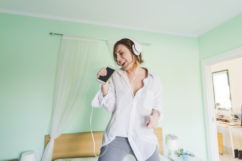 Junge Frau auf dem Bett, die Kopfhörer trägt und in ihr Smartphone singt, lizenzfreies Stockfoto