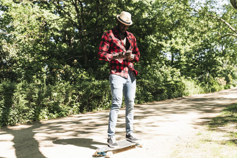 Cooler junger Mann, der im Park Skateboard fährt und sein Smartphone benutzt, lizenzfreies Stockfoto