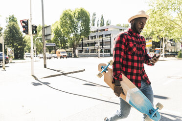 Cooler junger Mann mit Skateboard zu Fuß in der Stadt, mit Smartphone - UUF13850