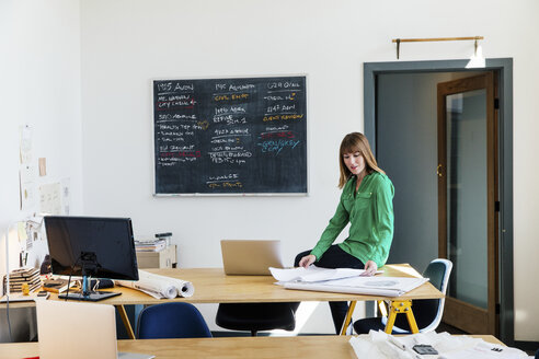 Frau sitzt auf einem Schreibtisch in einem Büro und sieht sich Baupläne an - ISF06124