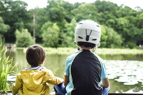 Zwei junge Brüder sitzen am Wasser, der ältere Bruder trägt einen Fahrradhelm, Rückansicht, lizenzfreies Stockfoto