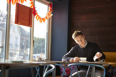 Young man alone in cafe drinking coffee and reading magazine - CUF13354