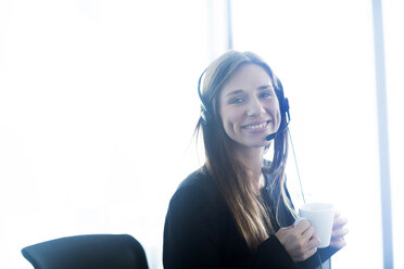 Young woman wearing telephone headset holding coffee cup looking at camera smiling - CUF13364