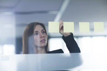 Blick durch ein Fenster auf eine junge Frau, die auf einen an das Glas geklebten Post-it-Zettel schreibt - CUF13368