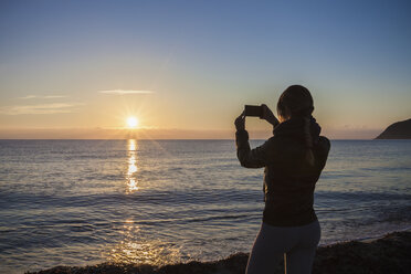 Silhouettenansicht einer jungen Frau, die den Sonnenuntergang über dem Meer fotografiert, Villasimius, Sardinien, Italien - CUF13391