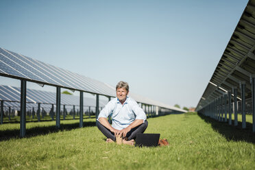 Mature man sitting barefoot on meadow, laptop and coffee to go cup, solar plant - MOEF01140